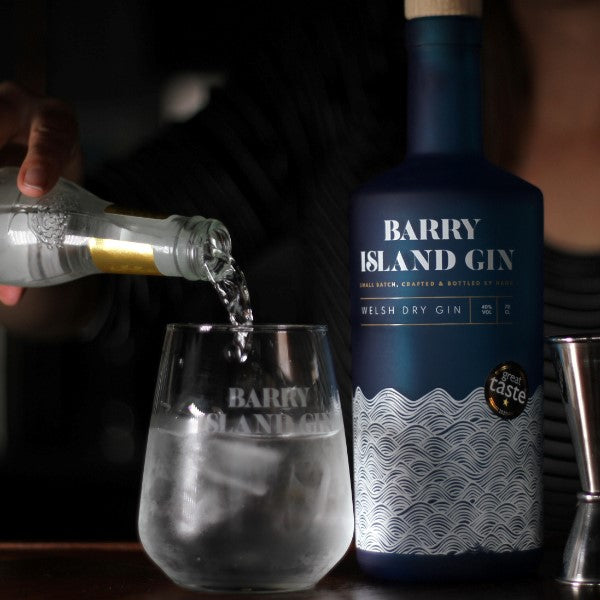 A Bottle of Barry Island Gin on a bar with a glass and a woman pouring tonic into the glass