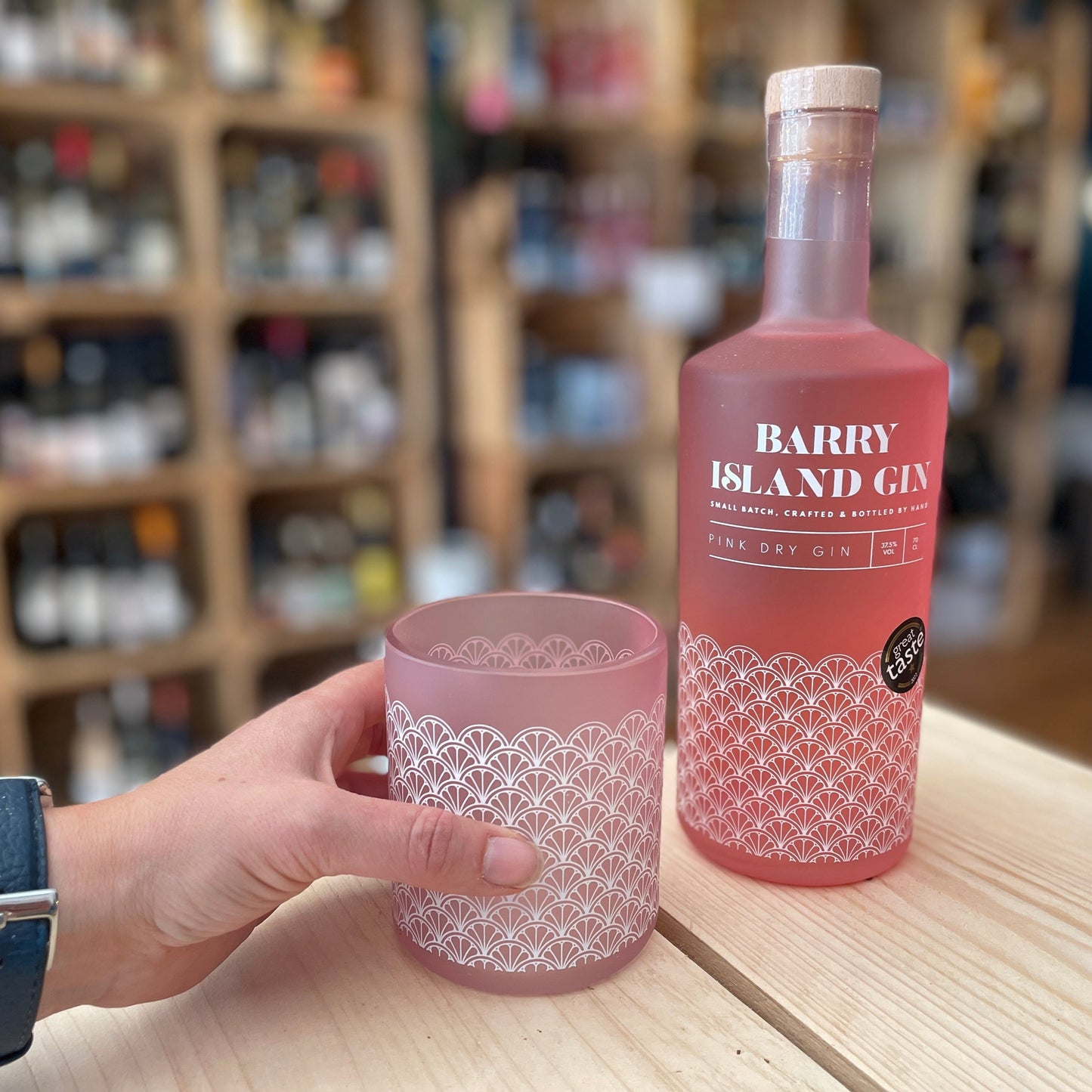 A hand holding a Barry Island pink gin glass on a table with a bottle of Barry Island pink gin next to it