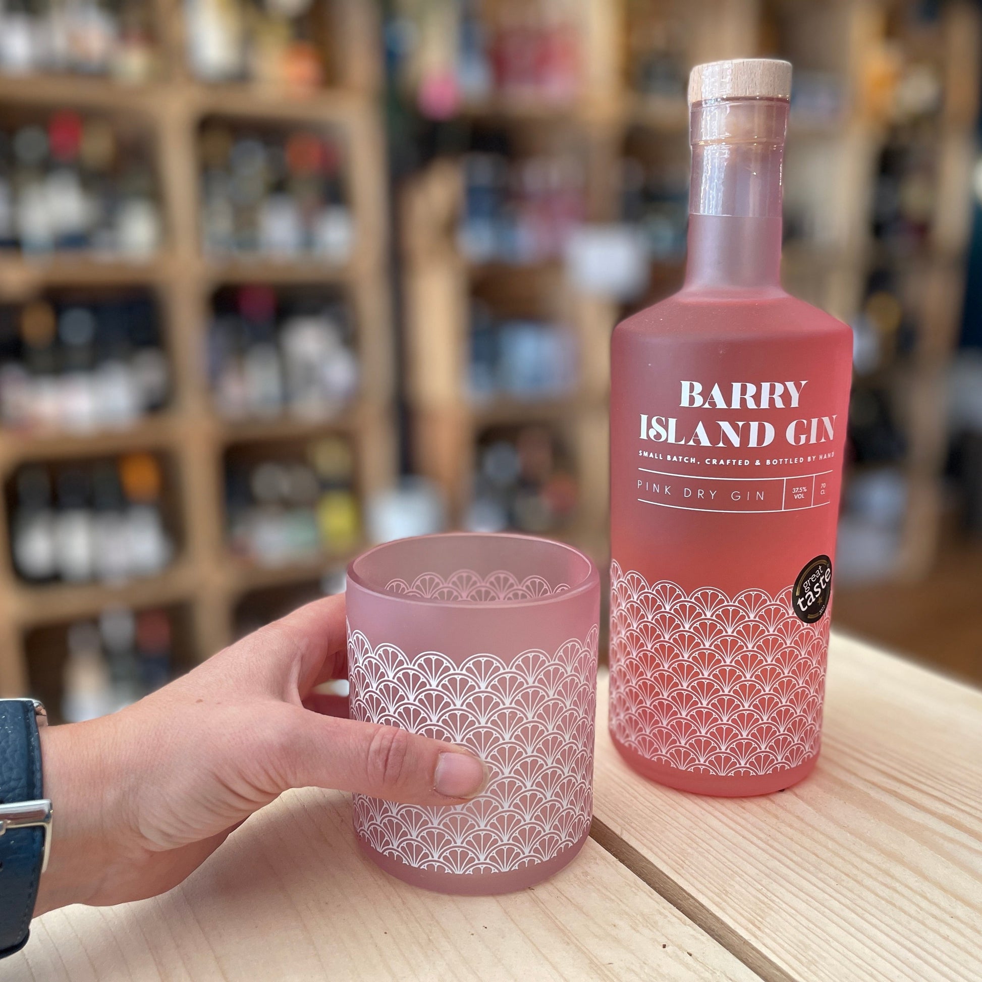 A hand holding a Barry Island pink gin glass on a table with a bottle of Barry Island pink gin next to it