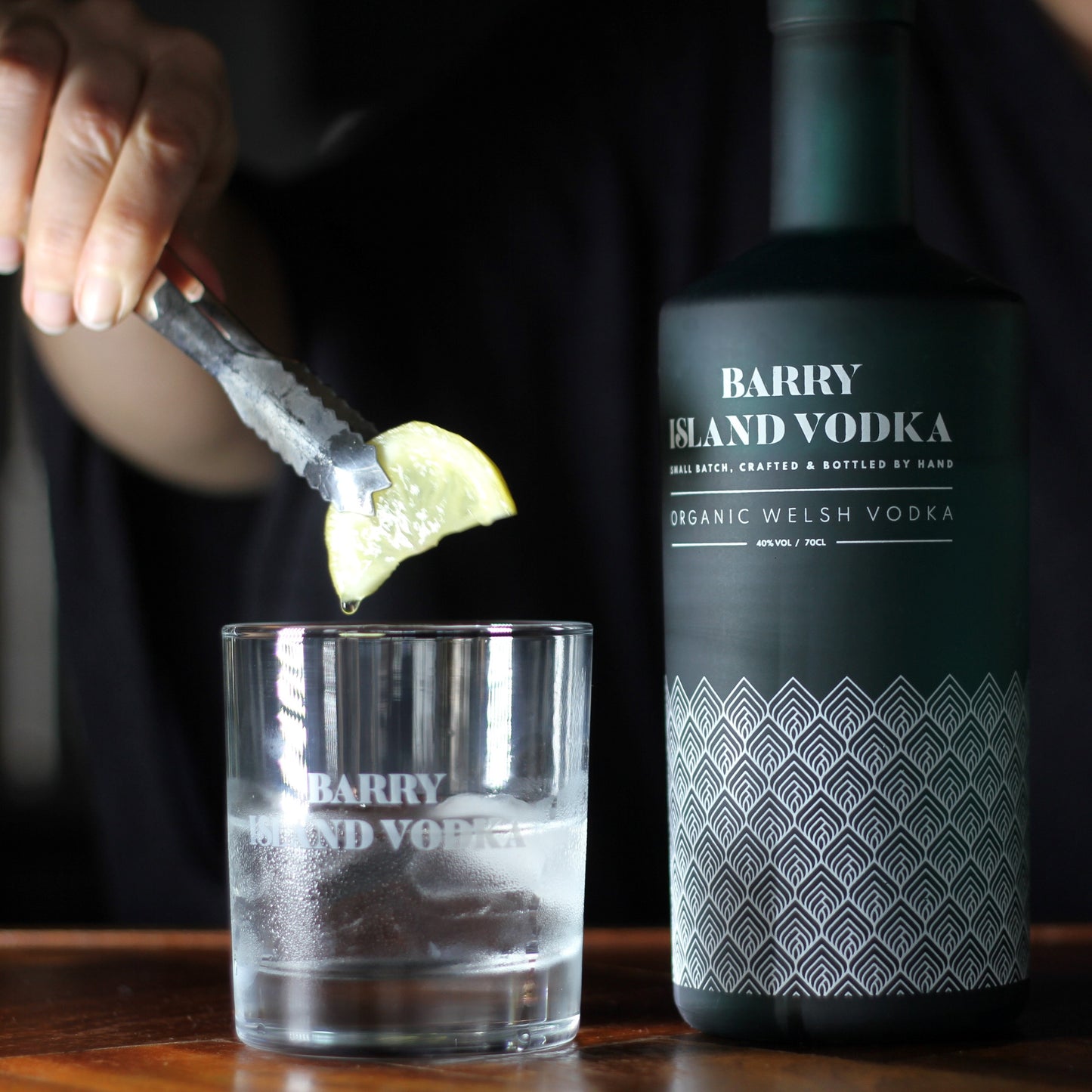 A bottle of Barry Island Vodka on a bar top with a glass with ice and vodka and a hand holding a lime
